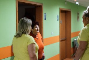 Senior woman engages in conversation with caregivers in a facility corridor.